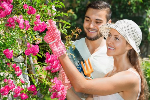 A man and woman are in a lush garden, surrounded by vibrant pink flowering bushes and green foliage. The woman, wearing a white sunhat and gardening gloves, is in the process of pruning or tending to the flowering shrub with a small garden tool. The man stands nearby, smiling and observing her work, dressed casually for outdoor activity. The garden features a mixture of well-maintained plants, dense flowering blooms, and leafy branches, with a background of trees providing natural shade. The scene is illuminated by natural daylight, suggesting clear weather, and the overall setting reflects an outdoor space suitable for gardening activities, aligned with professional lawn care and gardening services offered by Lawn Mowing East Ham in the local area around postcode E6, East Ham. This image highlights the care of flower beds, maintenance of garden shrubbery, and general outdoor landscaping work typical of a well-kept urban garden environment.