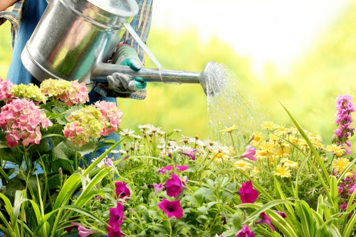 A person wearing gardening gloves and a blue jacket is using a metal watering can to water a vibrant flower bed in an outdoor garden. The flower bed contains a mixture of flowering plants, including pink, yellow, and purple blooms, with lush green foliage. The plants are arranged along a cultivated soil border, and some are spilling over a small wooden edging. In the background, blurred green trees and shrubs create a natural backdrop, indicating a well-maintained garden space typical of East Ham outdoor landscaping. The scene is illuminated by natural daylight, suggesting a clear, sunny day conducive to gardening activities. This setup exemplifies careful garden layout and plant care, consistent with professional gardening and lawn care services offered by Lawn Mowing East Ham in the local area.